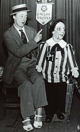 Photograph of a woman dressed in a man's trousers, jacket, tie, and straw boater and women's sandals sitting on a stool on a stage with curtains behind her; beside her are the striped blouse and dark trousers of a ventriloquist's doll draped over the edge of a large whicker basket; above the costume of the doll is the head of a woman made up to resemble a doll, her body presumably being concealed in the basket; on the curtains behind them is a banner with the words, Long Live The Queen, and it is possible that the entertainment is part of celebrations to mark the coronation of Queen Elizabeth II; the entertainers have been identified as Mrs. Waggot and Mrs. Short