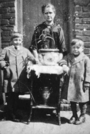 Photograph showing a woman dressed in a dark dress standing in front of the door to a house and behind a small table on which there are two vases; either side of the table,are two boys dressed in overcoats; one boy is aged approximately ten years,and the other,seven years; she has been identified as Mrs. Parker and Boys, of Haswell
