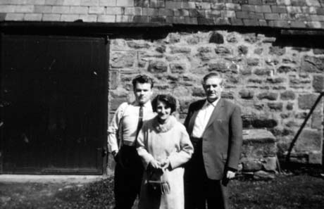 Photograph of a young man aged approximately twenty years, in shirt sleeves, standing outside a stone building; next to him is a middle-aged woman wearing a coat with a fur collar and carrying a handbag; next to her is a man wearing a suit and no tie; thay have been identified as Mr. and Mrs. Ivor James and Glyn