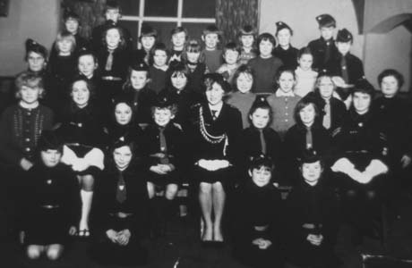 Photograph of twenty six girls in uniform and seven girls in ordinary clothes posed five rows in front of a window in a room; they are accompanied by two women in uniform; thay have been identified as members of the Girls' Brigade in Haswell