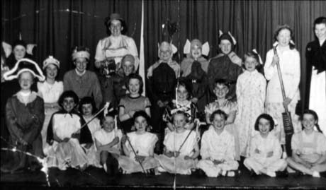 Photograph of twenty two children, aged between seven and ten years, in costume on stage with curtains behind them; five girls on the front row appear to be dressed as fairies and three children on the back row appear to be elves; two women in costume are also on the stage; they have been identified as Cubs and Brownies in Concert, Haswell
