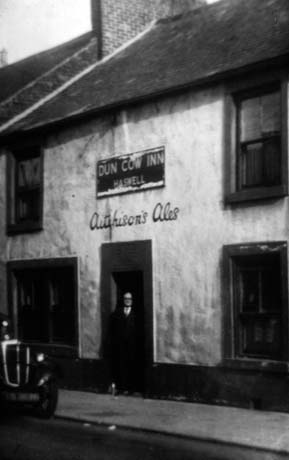Photograph of the facade of the Dun Cow Inn, Haswell, showing a plain stuccoed building with four sash windows and a plain doorway; the name of the public house is written on the wall as is Aitchison's Ales; a man is standing in the doorway of the building and part of the front of a car can be seen at the left of the photograph