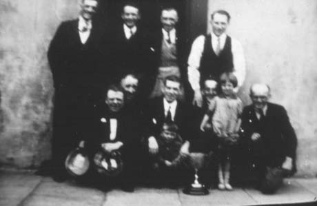Photograph showing four men, wearing suits, standing in the doorway of a building; in front of them are what appears to be five men, also wearing suits, kneeling down; a small girl, aged approximately four years, and a small boy aged approximately two years, are standing with the kneeling men; in front of the men is a trophy cup