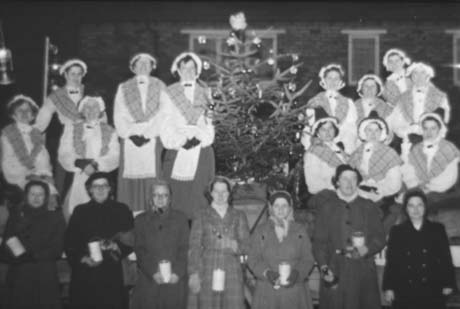 Photograph of eleven women, dressed in Welsh costumes, in a line, with a Christmas tree in the middle of them; in front of them are seven women dressed in overcoats and carrying collecting boxes; behind the women the facades of houses can be seen indistinctly; they have been identified as Haswell Women's Institute Carol Singers