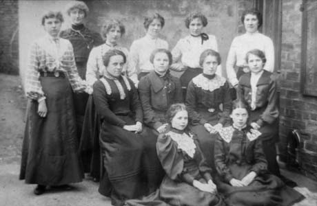 Photograph showing five young women in light-coloured blouses and dark skirts and one young woman in a dark blouse standing in front of a brick wall; in front of them are four young women in dark blouses and skirts sitting on chairs; and in front of them are two young women, dressed in a similar manner to those sitting, sitting on the floor; they have been identified as being in Haswell