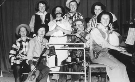 Photograph showing five women dressed as cowboys; one woman dressed as a bar tender; one woman dressed as a hillbilly and one woman dressed as a cowboy sitting at a piano; in front of the woman dressed as a hillbilly there is a frame on which bottles are hanging; they have been described as Haswell Women's Institute Hillbillies