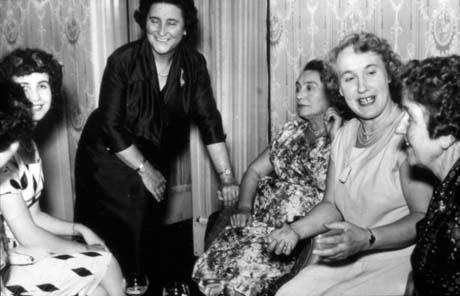 Photograph showing three women dressed in smart frocks sitting at the right of the picture with their backs to a wall, papered in an elaborate pattern; a woman in a dark frock is standing leaning slightly over and laughing in the centre of the picture; the lap and part of the face of a young woman can be seen on the left of the picture; they have been identified as attending a cocktail party for the Haswell Women's Institute