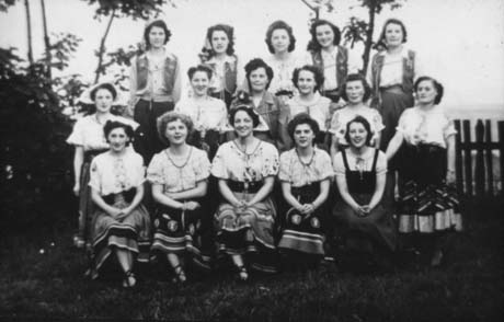 Photograph of sixteen women posed in three rows in front of trees; they are wearing full patterned skirts and frilly patterned blouses; thay have been described as taking part in a Gypsy Night arranged by Haswell Women's Institute