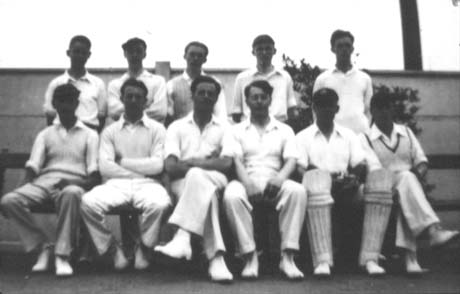 Photograph of eleven men in cricket whites posed in front of a wall in two rows; their faces are dark and cannot in most cases be seen; they have been identified as a cricket team in Haswell