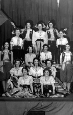 Photograph of fourteen women grouped in front of curtains, wearing patterned full skirts and patterned blouses and pantaloons and jerkins; they have been identified as members of Haswell Women's Institute on their Gypsy Night