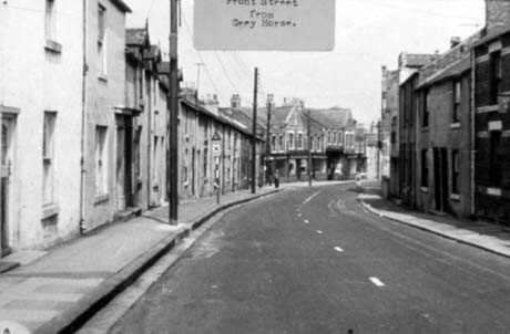 Photograph with a typed note reading: Front Street From Grey Horse, showing a road with terraced houses curving to the right; at the end of the terraced houses are a series of large buildings with shop fronts, which cannot be discerned; a Stop sign with a triangle on the top can be seen half way down the street; the figure of a man is at the far end of the street This photograph was submitted by Philip Soakell from the album 'History of Haswell and District', which was compiled by his grandfather Fred Soakell.