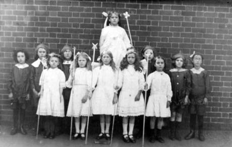 Photograph showing five girls, aged approximately seven years, standing in a row wearing white frilly dresses and carrying wands with stars at the top of them; behind them another girl of approximately the same age and also wearing a frilly dress and carrying a wand, is seated on some sort of eminence; at the side of the girl on the throne are six children dressed in dark frocks and white collars lined up on either side of her; they have been identified as being in Haswell