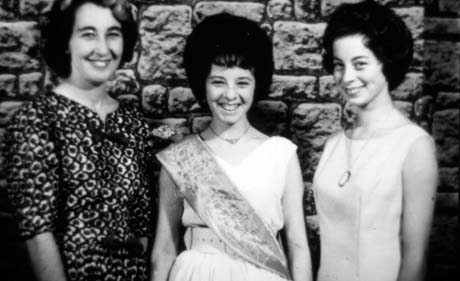 Photograph of a young woman wearing a light-coloured dress with a sash across her chest; on the right of the photograph is another young woman wearing a light-coloured dress and a pendant; on the left is woman wearing a patterned frock; the photograph has been described as Gala Queen, Haswell; they are standing against a stone wall