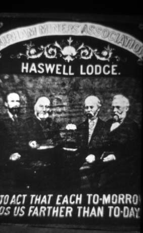 Photograph of a banner with the following words on it: Durham Miners Association Haswell Lodge To Act That Each Tomorrow Leads Us Farther Than To-Day; there are also the portraits of four men on the banner