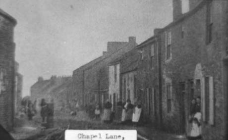 Photograph showing houses of different types and heights forming a continuous line curving to the left; indistinct figures can be seen in front of the houses, which are also indistinct in detail; they have been identified as Chapel Lane, Haswell This photograph was submitted by Philip Soakell from the album 'History of Haswell and District', which was compiled by his grandfather Fred Soakell.
