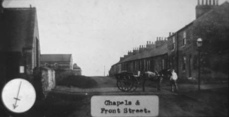 Photograph with a typed note reading: Chapels and Front Street Haswell, showing, on the left, the sides of two chapel buildings; on the right is a row of terraced houses; between the terrace and the chapels, are a horse harnessed to a two-wheeled trap and a man standing at the horse's head near a gas lamp This photograph was submitted by Philip Soakell from the album 'History of Haswell and District', which was compiled by his grandfather Fred Soakell.