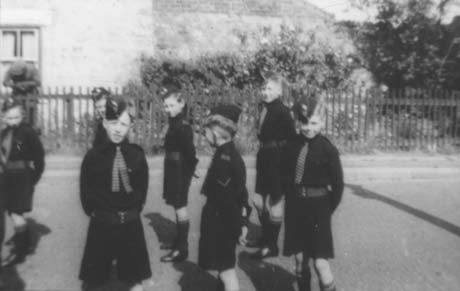 Photograph of eight boys, aged approximately eight years, standing in a road in twos, possibly waiting for a parade to begin; they are dressed in a uniform of dark shirt and shorts with a striped tie; across the road is a fence and part of the front of a house; a man can be seen leaning over the fence looking at the ground; the boys have been identified as members of the Church Lads' Brigade in Haswell