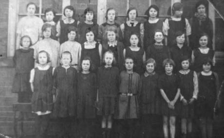 Photograph of twenty seven girls, aged approximately eleven years, posed in three rows against the windows of a brick building; thay have been identified as being in Haswell