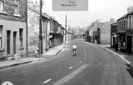 Photograph with a typed note reading: From Newsagents, showing a road running away from the camera and curving to the right in the distance; terraced houses can be seen on the left; shops, followed by terraced houses can be seen on the right; a cyclist is riding down the road away from the camera; the street has been identified as Front Street, Haswell; this photograph shows the same view as 0255, but from further away from the end of the street This photograph was submitted by Philip Soakell from the album 'History of Haswell and District', which was compiled by his grandfather Fred Soakell.