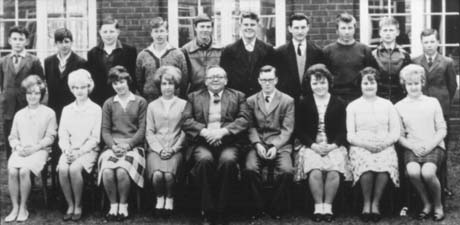 Photograph of eleven boys and seven girls, aged approximately fifteen years, posed in two rows in front of a brick building; a man is sitting in the middle of the front row; they have been identified as pupils at Haswell and Shotton Senior School