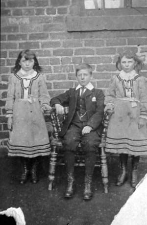 Photograph of a boy, aged approximately twelve years, dressed in a suit, waistcoat, stiff collar, and boots, sitting on a Windsor chair; on the left is a girl, aged approximately ten years, dressed in a frock with coloured bands round its bodice, cuffs and hem, and boots; on the right is a girl aged approximately eight years, wearing a dress exactly the same as that worn by the other girl; the photograph ahs been identified as Mrs. Hardy, Haswell