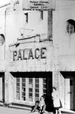 Photograph with a typed note reading Palace Cinema, Haswell. Opened 1912. Closed and Demolished 1962, showing the facade of the cinema in a dilapidated state with cracked paintwork; the word Palace can be seen on it; a woman wearing an apron and the legs of a man sitting on the sill of a window with a small child on his knee can be seen This photograph was submitted by Philip Soakell from the album 'History of Haswell and District', which was compiled by his grandfather Fred Soakell.
