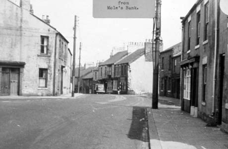 Photograph with a typed note reading: from Mole's Bank, showing a road curving round to the left with another road coming into it from the left;on the corner of the two roads is a house; on the right side of the picture are shops and houses; a car can be seen driving away from the camera in the distance; the photograph has been identified as showing Front street, Haswell, from Moles Bank This photograph was submitted by Philip Soakell from the album 'History of Haswell and District', which was compiled by his grandfather Fred Soakell.