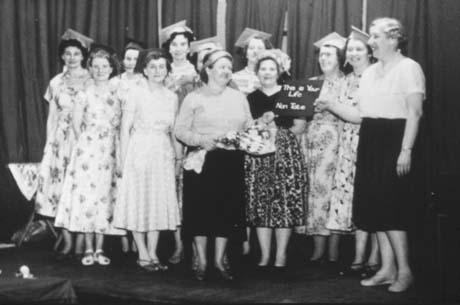 Photograph showing twelve women, wearing summer dresses, standing on a stage in front of curtains; six of them appear to be wearing mortar boards; a woman in the middle of the group is holding a bouquet of flowers and the woman next to her is holding a notice reading: This Is Your Life Nan Tait; they have been identified as members of Haswell Women's Institute