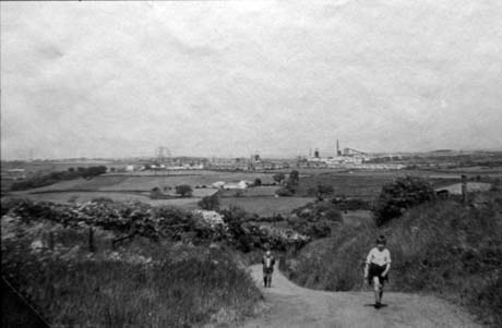 Photograph showing a road in the forefront of the picture running towards the camera; two people can be seen walking towards the camera; behind the road are open fields and beyond them, on the horizon, houses and three winding gear of collieries; the photograph has been described as Back Loden Road, Haswell This photograph was submitted by Philip Soakell from the album 'History of Haswell and District', which was compiled by his grandfather Fred Soakell.