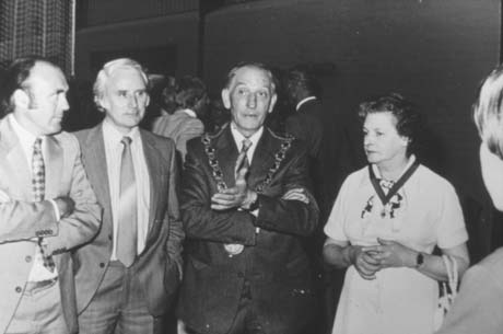 Photograph showing a man, wearing a suit and tie and a chain of office, standing with two men on the left, and with a woman on the right, wearing a dress and a ribbon with a medallion of office on it; indistinct figures can be seen in the background; the man in the chain has been identified as Councillor A. McAteer and the woman as Mrs. Margaret McAteer