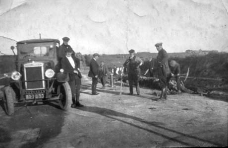 Photograph showing a motor car, at the left of the picture, facing towards the camera; its registration is BR7835; two men are leaning against the car; in the centre of the picture are seven men in work clothes of suits, waistcoats and caps, with picks in their hands digging a hole in the road; standing near them is a man who appears to be wearing a cap and gaiters and who may be the chauffeur of the car; beyond the road is open country; the men have been identified as Albert and Jack Conroy and Jack Cornforth, and the scene as road works at Haswell