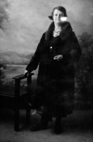 Photograph showing a full-length portrait of a woman wearing a dark coat; she has short hair and is wearing a necklace; she is standing, and resting her right hand on a small table, in a photographer's studio; she has been identified as Mrs. Anderson of Haswell