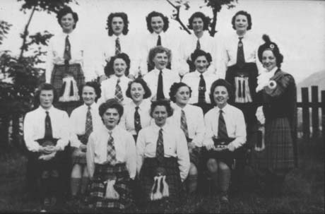 Photograph of fifteen women, posed in four rows, wearing blouses, tartan ties, kilts and sporrans; a woman is standing on the right of the group wearing a Highland bonnet, a tartan over her shoulder, and a frilly blouse, presumably the clan chief; the women have been identified as members of Haswell Women's Institute during a Scottish night