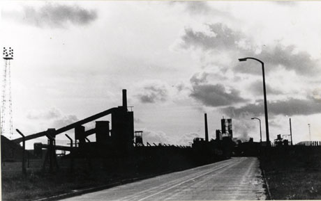 Photograph showing the surface of a metalled road running away from the camera with the buildings of a colliery on the left-hand side; the details of the buildings cannot be seen as they are outlined against the sky; the photograph is described as New Hawthorn Shaft