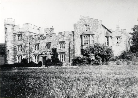 Photograph of the exterior of the front of Hawthorn Towers, Hawthorn; the photograph shows a castellated building with Gothic windows, a large porch and a coat of arms above the porch; the photograph is taken across the grass in front of the house