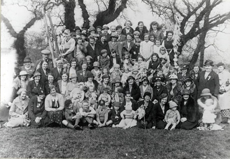 Photograph of approximately ninety people posed in front of trees; they are comparatively formally dressed, wearing overcoats, dresses, suits and hats; a woman on the front row is holding a Union Jack; the photograph has been identified as marking the celebrations in Hawthorn of the Silver Jubilee of the reign of George V
