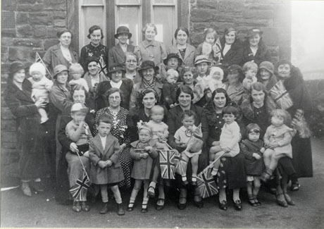 Photograph of approximately fifty women and children posed outside a stone building; they are formally dressed in overcoats, dresses, and hats; ten of the children are carrying Union Jacks; the photograph has been identified only as Celebrations at Hawthorn, but it is possible that it, like hawt0011 and hawt0012, marks the Silver Jubilee of the reign of King George V