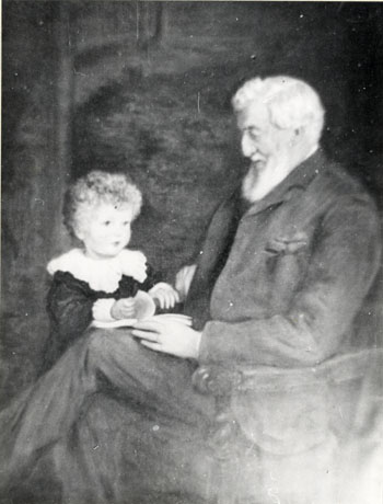 Photograph of an elderly man, with white hair and beard, shown from his left-hand side sitting with his legs crossed on a wooden chair with arms; at his right knee, a small boy with short curly hair and wearing a garment with lace collar and cuffs, is standing turning the pages of a book resting on the elderly man's knee; the man and boy have been identified as Richard Lawrence Pemberton and his grandson, R.L. Stapylton Pemberton, Hawthorn Towers, May 1893