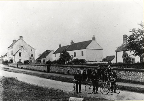 Photograph showing a narrow road with a wall beyond it, and, beyond the wall, seven houses set back from the road; the houses appear to be of at least nineteenth century origin; on the side of the road in front of the wall a group of four men, one woman and two children are standing looking at three men with bicycles who are standing in the road; the photograph has been identified as depicting a scene in Hawthorn