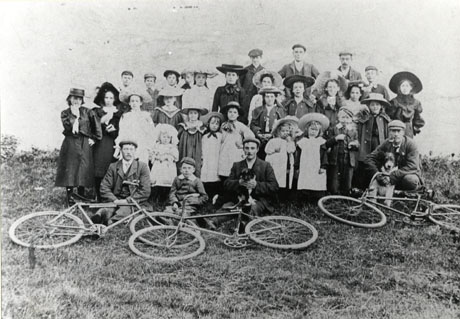 Photograph of a group of twenty six children, described as children from Hawthorn, with three men; they are all posed behind another three men, who are squatting on the ground behind three bicycles, which are lying on the ground; all the children are smartly dressed; all three men with the bicycles are formally dressed in suit, tie and cap and are smoking pipes; two of these men are each holding a dog