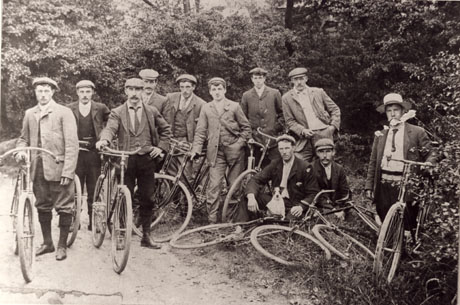 Photograph of eleven men with bicycles posed against a background of trees;the men are dismounted and are holding their bicycles; all are dressed in suits and are wearing bicycle clips or have the legs of their trousers tucked into their socks; one man is holding a dog; the men have been identified as members of the Hawthorn Cycling Club