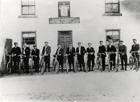 Photograph of twelve men standing in a row, each holding a bicycle, outside The Stapylton Arms public house; the facade of the public house may be seen; above the door is a notice reading Alice Johnson Licensed to Retail Ales, Stout, Wines, Spirits & Tobacco; the men are formally dressed and have been identified as members of the Hawthorn Cycling Club