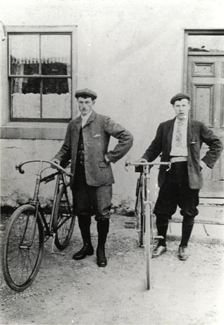 Photograph of two men standing in front of The Stapylton Arms, each with a bicycle; a window and part of the door of the public house may be seen; the men are formally dressed and have been identified as Luke Lamb and Fred Bennison