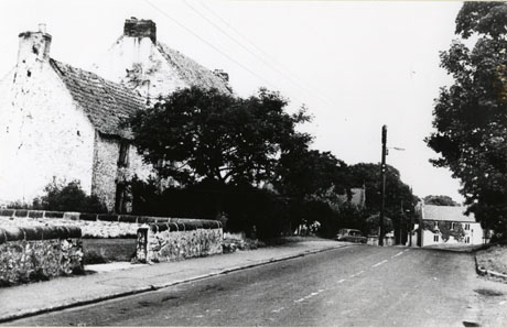Photograph of houses, mainly obscured by trees, on one side of a road, the surface of which is in the foreground and running away to the right, of the photograph; the houses have tiled roofs which appear to be of some age; the facade of another house can be seen in the distance on the opposite side of the road from the other houses; a car is parked in the middle distance; the photograph has been identified as Hawthorn Village