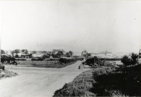 Photograph showing the meeting of four narrow roads running through fields; hedges and fences lining the roads can be seen, as can very small indistinct figures at the side of the left-hand road and approaching up the road leading towards the camera; the photograph has been identified as The Crossroads on the Old A19 Road at Hawthorn Village