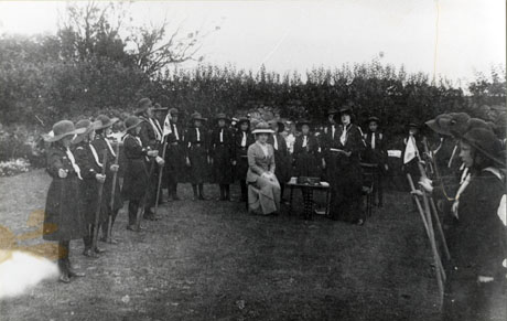 Photograph of approximately twenty five Girl Guides in uniform drawn up in three sides of a square on grass surrounded by a rough stone wall; within the square a small table can be seen with a Guider standing beside it and a woman in a long skirt, fitted jacket and large hat seated beside it; the Guides have been identified as the !st Hawthorn Guides, the Guider as Miss Pemberton of Hawthorn Towers, and the woman in the hat as the Hon. Agnes Ridley; the photograph was taken on 5 August 1914