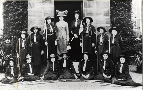 Photograph of thirteen Girl Guides in uniform posed in front of what may be the porch of a large house with a Guider and a woman in a long skirt, fitted jacket and large hat, in the centre of the group; the Guides have been identified as the !st Hawthorn Guides and the Guider as Miss Pemberton of Hawthorn Towers and the woman in the hat as the Hon. Agnes Ridley of Ravensworth Castle; the photograph was taken on 5 August 1914