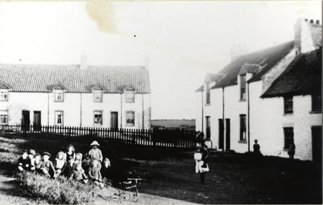 Photograph showing a row of terraced houses in the middle distance and another row on the right-hand side of the photograph at right angle to the other row; in front of the first row is a fence and in front of the fence is an open area of grass; at the front of the picture an indistinct group of thirteen children with a tricycle can be seen; the indistinct figures of four other children can be seen; the photograph has been identified as Village Green, Hawthorn