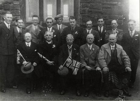 Photograph of fifteen men posed in front of a stone building; the men are dressed in overcoats and have rosettes in their lapels; the men on the front row are holding Union Jacks; one of the men on the front row is a clergyman wearing a dog collar; the photograph has been identified as depicting celebrations in Hawthorn to mark the Silver Jubilee of the reign of King George V; the men have been identified as follows: back row, left to right: Joe Irving; Mr. Patterson; Chas. Newby; Bill Butt; Bob Reed; Gerald Scully; Wilf Tate; Bob Turner; Bob Matthews; Ralph Turner; front row: Jos. Tate; Rev. Widdes; Luke Lamb; Geo. Cresswell; Grandad Newby
