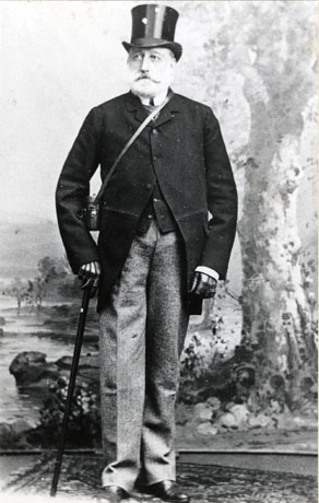 Photograph, full-length, taken in a photographer's studio; of a middle-aged man with a moustache and beard and wearing a top hat, leather gloves, a black jacket and waistcoat, and lighter coloured trousers; he is holding a cane and carrying binoculars in a case; it is possible that he is dressed for a day at the races; he has been identified as Major Stapylton